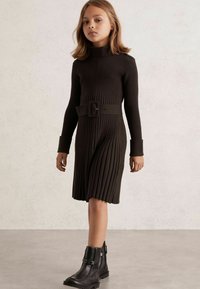 Bruine geribde jurk met een hoge hals, een geknoopte taille en lange mouwen. Gecombineerd met zwarte enkellaarsjes met decoratieve accenten.