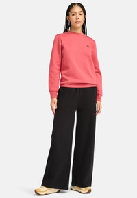 Sudadera de color coral con cuello redondo y logo, combinada con pantalones de pierna ancha negros y zapatillas beige, con una tela texturizada y suave.