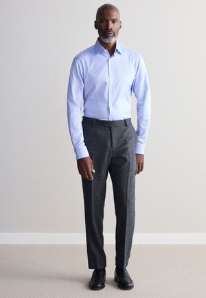 Un uomo è in piedi davanti a uno sfondo uniforme, indossa una camicia azzurro chiaro con bottoni, pantaloni eleganti grigio scuro e scarpe eleganti nere.