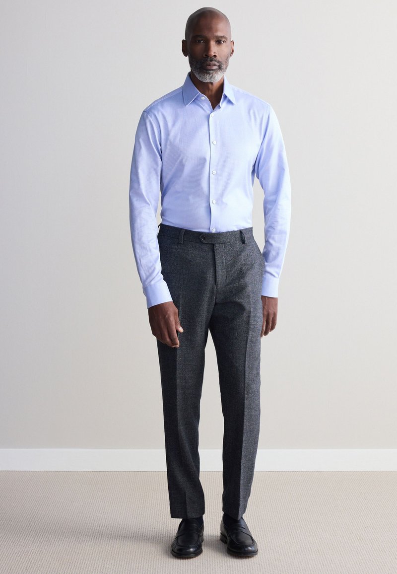 Un homme se tient devant un fond uni portant une chemise bleu clair à boutons, un pantalon de costume gris foncé et des chaussures de ville noires.