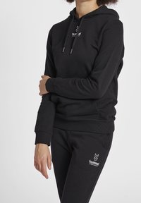 Felpa nera con cappuccio e pantaloni coordinati in misto cotone. Cappuccio concordabile con lacci di regolazione, polsini a coste e logo ricamato sul petto.