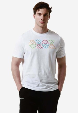 T-shirt in cotone bianco con un design del logo multicolore sul petto, scollo rotondo e maniche corte. Il modello indossa pantaloni neri.