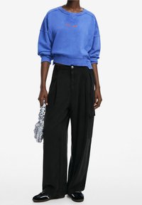 Felpa blu corta con testo rosso, abbinata a pantaloni neri a gamba larga. Accessoriata con una borsa argentata sgualcita e sneakers nere.