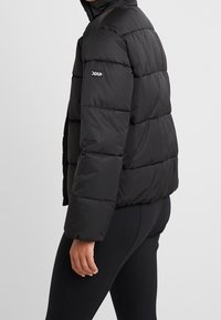 Giacca a vento nera con cappuccio, caratterizzata da un design trapuntato, polsini elasticizzati e un logo sulla manica; texture liscia e lucida.