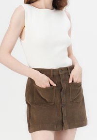 Femme portant un haut blanc côtelé sans manches rentré dans une jupe en velours côtelé marron à boutons sur le devant, avec de grandes poches avant dans lesquelles elle a les mains.