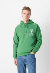 Πράσινη φούτερ με καγκουρό τσέπη και κουκούλα με κορδόνι. Έχει λευκή γραφιστική εκτύπωση στο στήθος. Συνδυάζεται με μπλε τζιν παντελόνι.