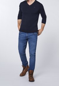 Marinblå v-ringad tröja med stickad textur, kombinerad med blå jeans. Bruna snörstövlar med metallhål. Modellen står med händerna i fickorna.