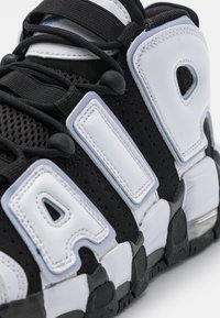 Fekete sportcipő, amely hálós és bőr anyagok keverékéből készült, kiemelkedő fehér logóval, texturált felülettel és hangsúlyos fűzőrendszerrel rendelkezik.