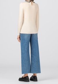 Kvinna iklädd en krämfärgad långärmad topp, vida blå jeans med fransade fållar och svarta klumpiga loafers, stående vänd bort från kameran mot en enfärgad bakgrund.