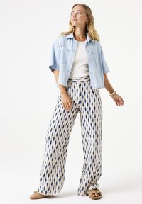 Camisa de mezclilla clara de manga corta sobre una camiseta blanca; pantalones anchos con patrón en beige y azul marino con formas en gota; sandalias de color arena.
