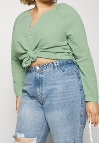 Blusa a maniche lunghe verde menta con fronte annodato, abbinata a pantaloni corti in denim blu a vita alta e strappati. Tessuto testurizzato.