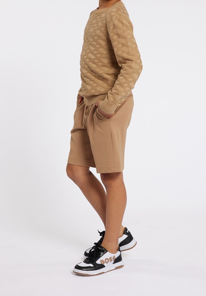 Enfant portant un pull texturé beige, un short beige assorti et des baskets noir et blanc avec le logo "BOSS", debout les mains dans les poches.
