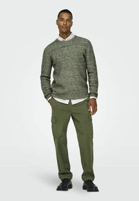 Groene gebreide trui met een gestructureerde afwerking, gedragen over een witte blouse met kraag, gecombineerd met olijfgroene cargobroeken en zwarte schoenen.