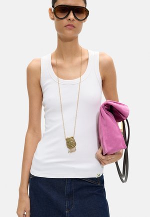 Kobieta w dużych ciemnych okularach przeciwsłonecznych, białym topie, ciemnej dżinsowej spódnicy, długim złotym naszyjniku z zawieszką, trzyma złożoną różową zamszową torbę z ciemnym paskiem.