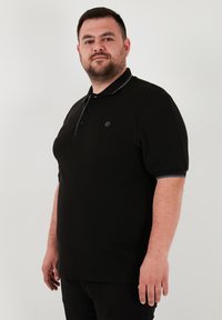 Čierne polo tričko s krátkymi rukávmi, ktoré má golier s kontrastným modrým lemovaním a malým logom na hrudi. Zloženie z bavlny.
