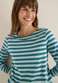 Smilende kvinde med brunt hår iført en teal og lyseblå stribet langærmet skjorte, stående mod en lysfarvet væg.