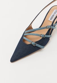 Tumman sinisen denim-kankaan kärkitossut, joissa on ristiin menevät vaaleansiniset remmit, hopeiset soljet ja sileä beige sisäpohja.