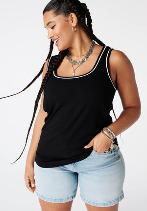 Vrouw met lang gevlochten haar die lacht, draagt een zwart tanktop met witte bies, lichte spijkershorts en gouden en zilveren sieraden.