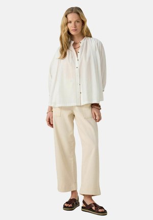 Blouse blanche à manches longues avec un col rond et une fermeture à boutons, associée à un pantalon large crème et des sandales à lanières marron.