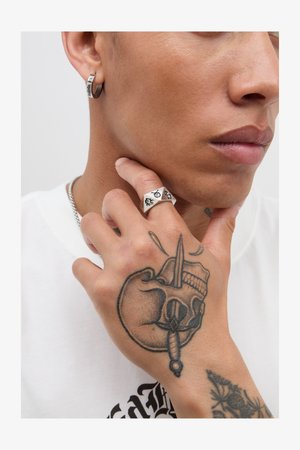 Ezüst karika fülbevaló és egy vaskos ezüst gyűrű fekete grafikai mintákkal, egy részletes tetoválásokkal díszített kézen, semleges háttér előtt.