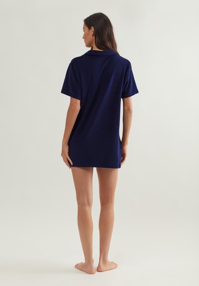 Γυναίκα στέκεται ξυπόλητη με την πλάτη γυρισμένη, φορώντας ένα μπλε ναυτικό φόρεμα από πετσέτα με κοντά μανίκια σε ένα απλό ανοιχτό φόντο.