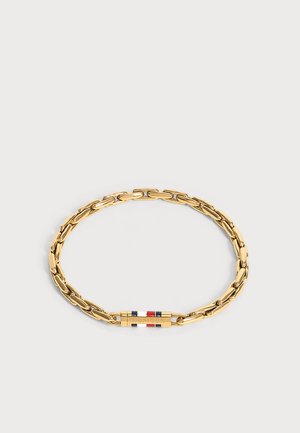 Bracelet chaîne en or avec un fermoir rectangulaire présentant des rayures rouges, blanches et bleues et le nom de la marque gravé sur un fond clair uni.