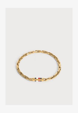 Bracelet chaîne en or avec un fermoir rectangulaire présentant des rayures rouges, blanches et bleues et le nom de la marque gravé sur un fond clair uni.