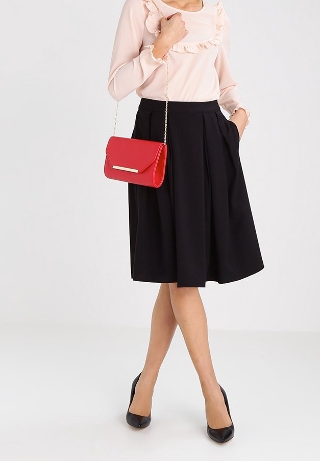 Femme portant un chemisier rose pâle à volants, une jupe noire plissée, des talons noirs, et tenant un sac rouge avec une bandoulière en chaîne dorée.