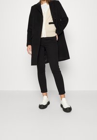 Abrigo negro de diseño largo, cárdigan crema con un bolsillo, vaqueros negros y zapatillas blancas con suela negra. Textura suave en toda la prenda.