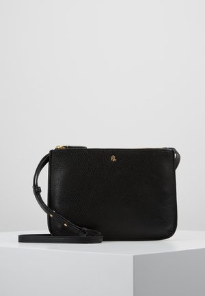Sac bandoulière en cuir noir texturé avec sangle réglable et petit logo doré à l'avant, posé sur une surface blanche sur fond gris.