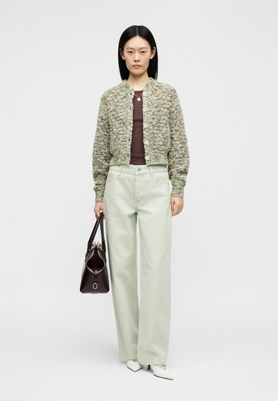 Femme portant un cardigan texturé boutonné, un haut bordeaux, un pantalon clair à jambes larges, des talons blancs, tenant un sac à main marron foncé, debout devant un fond uni.