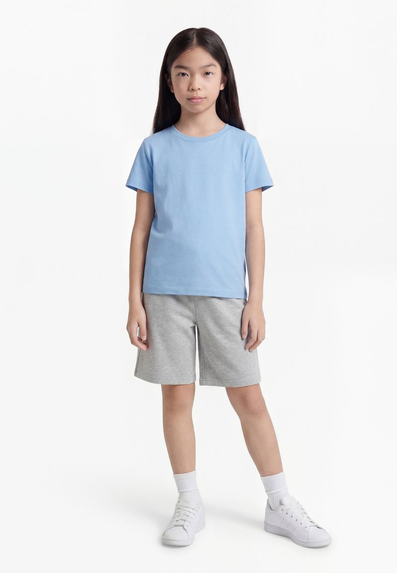 Fetiță stând cu fața înainte, purtând un tricou albastru deschis, pantaloni scurți gri, șosete albe și adidași albi pe un fundal alb simplu.