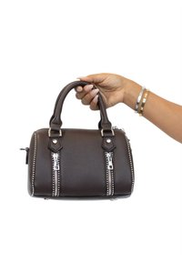 Bolso de cuero marrón con dos detalles de cremallera plateados, forma estructurada, asas superiores y superficie texturizada. Sostenido a mano con pulseras visibles.