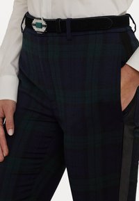 Los pantalones a cuadros en tonos azul marino y verde cuentan con paneles laterales negros, bolsillos delanteros y un cinturón negro elegante con un cierre decorativo plateado y un acento verde.