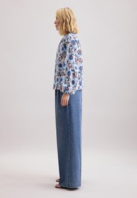 Giacca con stampa floreale nei toni di blu, rosso e bianco, abbinata a una gonna di jeans blu a vita alta. Modella in profilo, indossa sandali.