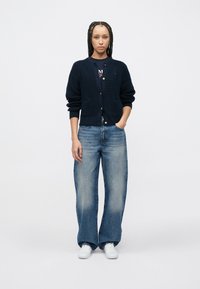 Πλεκτή μπλε ναυτική ζακέτα με δύο μπροστινές τσέπες και κλείσιμο με κουμπιά; συνδυασμένη με ανοιχτό μπλε τζιν παντελόνι με φαρδιά πόδια και λευκά αθλητικά παπούτσια.