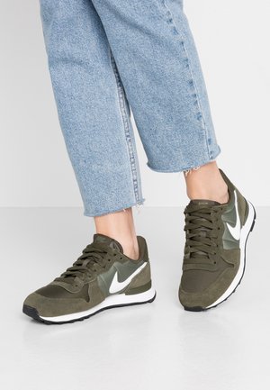 Pieds chaussés de baskets Nike vert olive avec un swoosh blanc, assortis à un jean court bleu clair effiloché, debout sur un sol blanc lisse.