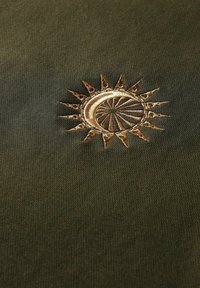 Olivgrüner Stoff mit komplizierten goldenen Sonnenstickereien, die scharfe Strahlen und ein kreisförmiges Zentrum zeigen, mit einer glatten Textur.