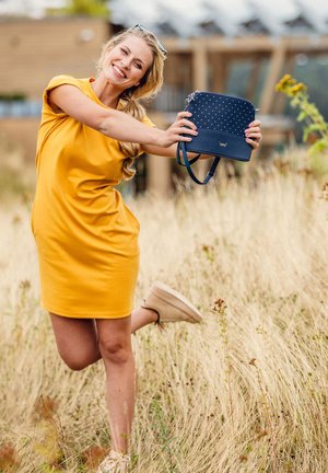 Navy handbag with polka dot pattern, textured leather, and logo. Model wears a yellow dress and light shoes, standing in tall grass.