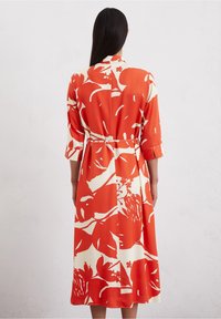 Donna con lunghi capelli scuri che indossa un vestito midi con una grande stampa floreale rossa e bianca, maniche a tre quarti e una cinta in vita, vista di spalle.