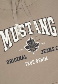Kaptur hoodie w jasnym beżu z wyraźnym, ciemnoniebieskim napisem "MUSTANG". Posiada grafikę liścia i nadrukowane logo w białych akcentach. Miękka tekstura.