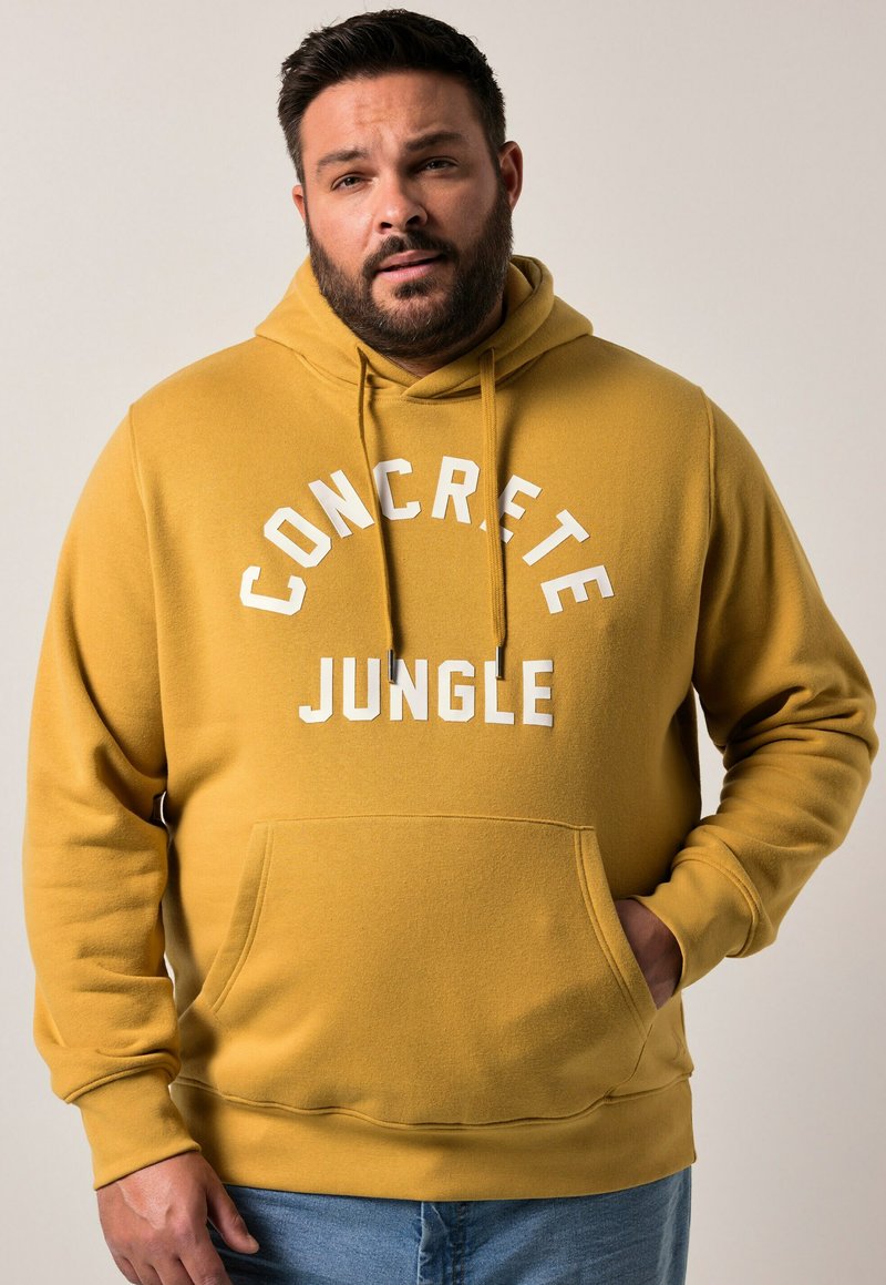Žlutá mikina s kapucí se nápisem "CONCRETE JUNGLE" v bílém písmu, s přední kapsou a pružnými manžetami. Materiál vypadá měkce.