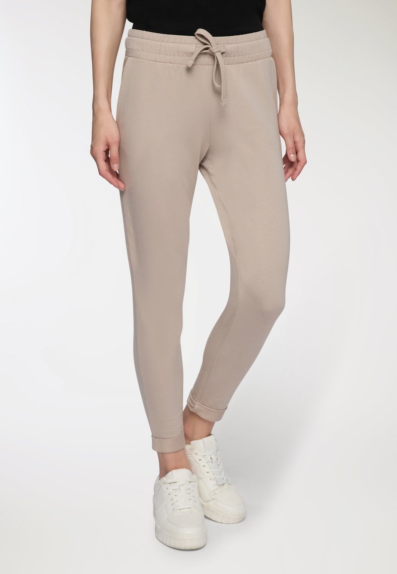 Beżowe spodnie dresowe z elastycznym pasem, sznurkiem i tapered nogawkami, zestawione z białymi sneakersami. Gładka tekstura, swobodny design.