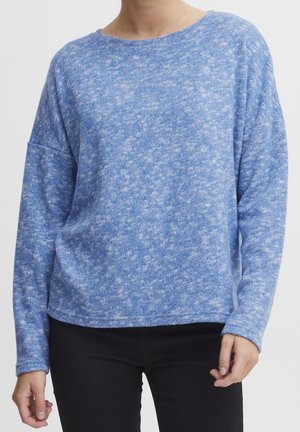 Osoba ubrana w miękki, długi rękaw, niebieski sweter w cętki, zestawiony z czarnymi spodniami, pokazana od szyi do bioder.