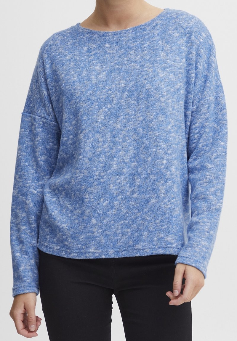 Personne portant un pull doux à manches longues bleu tacheté associé à un pantalon noir, montré du cou jusqu'aux hanches.