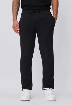 Homme portant un pantalon noir à cordon avec poches et des baskets blanches, se tenant avec les mains détendues sur les côtés.