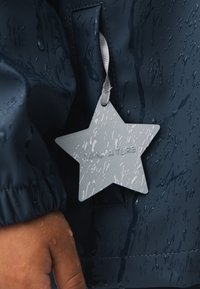 Marineblauer wasserdichter Mantel mit Wassertropfen. Verfügt über ein silbernes, sterneförmiges Etikett mit der Aufschrift "MINIWATURE", das an einem grauen Band befestigt ist.