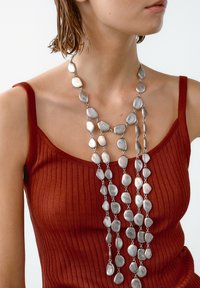 Silberne Metallkette mit mehreren eiförmigen Gliedern, geschichtetem Design und polierter Oberfläche. Getragen über einem rostfarbenen, gerippten Tanktop.