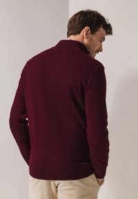 Bordeauxroter Pullover mit hohem Kragen, gerippten Bündchen und Saum. Hergestellt aus weichem Stoff, zeichnet er sich durch eine glatte Textur und eine figurbetonte Passform aus.