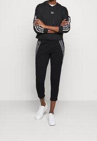 Černá zkrácená mikina s bílými logy na rukávech, v kombinaci s černými kalhotami se bočními pruhy, doplněna bílými teniskami.